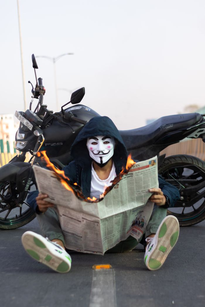 Home A person in a Guy Fawkes mask sits on the street with a burning newspaper, near a motorcycle.
